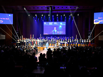 Blick aus dem Zuschauerraum auf die Bühne des Mozartsaals. Dort im Hintergrund das SAP Sinfonieorchester sitzend, auf einem roten Teppich schreitet ein Absolvent richtung Bühne, der auf einer Leinwand von vor zu sehen ist. 