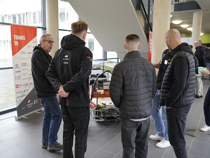 Fahrsimulator im Hintergrund, im Vordergrund 4 Menschen im Gespräch