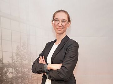 Porträt einer lächelnden Dame mit hellen Haaren, großer Brille und schwarzem Blazer vor einer Fotowand der DHBW Mannheim, auf der man ganz blass den Campus sehen kann. 