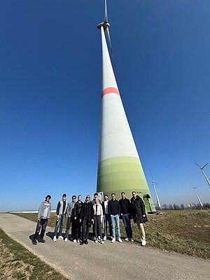 Gruppe mit 11 Studierenden vor einem Windrad - von unten fotografiert, sodass man das Windrad komplett sieht sowie den strahlend blauen Himmel. 