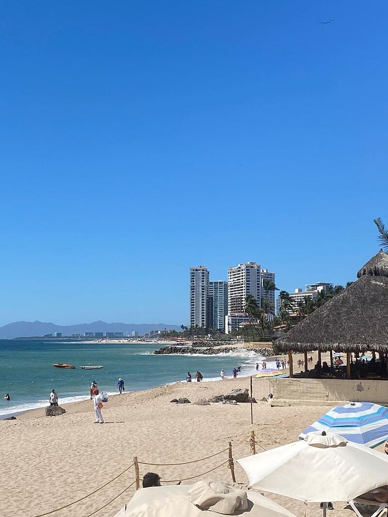 Strand mit Schirmen im Vordergrund, wenigen Menschen und mehrstöckigen Häusern im Hintergrund.