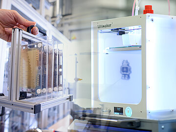 Collage aus zwei Bildern: Links hält eine Hand eine Halterung mit mehreren hintereinander gereihten Brennstoffzellen, rechts sieht man einen 3D-Drucker.