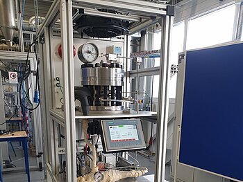 Der elektrochemische Verdichter: Ein vertikaler Versuchsaufbau in einem Aluminium-Profilrahmen in einer Laborhalle. Das Gerät besteht aus mehreren Ebenen: Oben befindet sich ein zylindrischer Druckbehälter mit Manometer, in der Mitte ein Siemens-Touch-Bedienpanel und darunter verschiedene Rohrleitungen, Pumpen sowie elektronische Steuerungskomponenten. Rechts daneben steht ein weißes Kühlgerät.