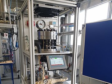 Der elektrochemische Verdichter: Ein vertikaler Versuchsaufbau in einem Aluminium-Profilrahmen in einer Laborhalle. Das Gerät besteht aus mehreren Ebenen: Oben befindet sich ein zylindrischer Druckbehälter mit Manometer, in der Mitte ein Siemens-Touch-Bedienpanel und darunter verschiedene Rohrleitungen, Pumpen sowie elektronische Steuerungskomponenten. Rechts daneben steht ein weißes Kühlgerät.