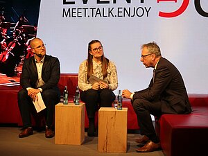 Links zwei Studierende auf einer roten Couch sitzen dem Oberbürgermeister auf einer rechten Couch gegenüber und hören ihm zu.