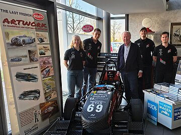 1 Studentin, 3 Studenten und ein älterer Herr stehen um einen schwarzen Rennwagen herum; links eine Werbewand von Bugatti und rechts davon Buchaufsteller. 