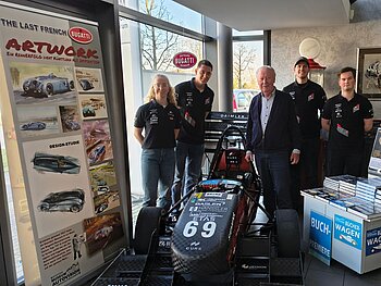 1 Studentin, 3 Studenten und ein älterer Herr stehen um einen schwarzen Rennwagen herum; links eine Werbewand von Bugatti und rechts davon Buchaufsteller. 