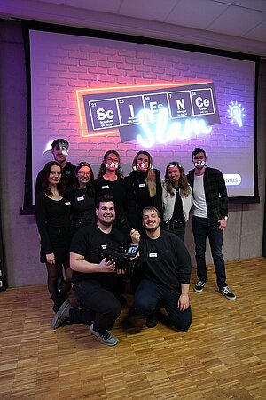 Gruppenfoto vor einer Präsentation, auf der die Worte Science Slam in Form von Scrabble-Steinen dargestellt wird. 