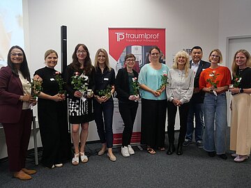 Gruppenbild mit 8 Professorinnen mit Blumen in der Hand und 1 Professor vor einem Aufsteller mit dem Logo des Programms Traumberuf Professorin Plus. 