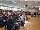 Großer Hörsaal mit zahlreichen Studierenden, die auf Sitzreihen einen Vortrag verfolgen. Vorne rechts befindet sich ein Rednerpult und eine Person, die zum Publikum spricht.
