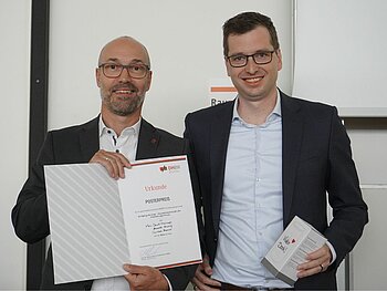 Porträt von zwei lachenden Professoren mit schwarzen Anzügen. Der linke eine Urkunde in der Hand. 