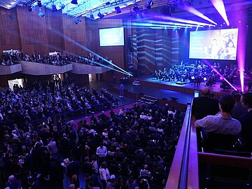 Blick von der Empore auf die Bühne. Der Raum ist blau-lila beleuchtet, die Sitzplätze alle belegt, auf der Bühne spielt das Orchester, auf der Leinwand sieht man einen Filmausschnitt. 