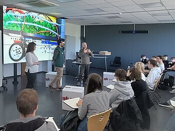 Gruppe von Schüler*innen an Stühlen in einem Halbkreis sitzend. Vor ihnen eine Professorin und zwei Professoren, die vor einer deckenhohen digitalen Leinwand stehen, auf der der Wasserstoffbus der Schüler*innen zu sehen ist. 