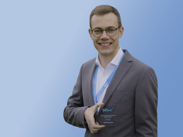 Porträt eines strahlenden jungen Mannes im Anzug, der seinen Glas-Award mit blauer Schrift in der Hand hält, vor blauem Hintergrund. 