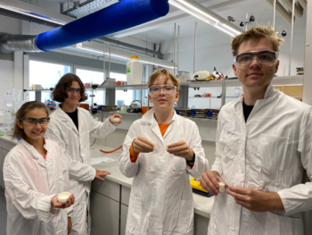 4 Jugendliche mit Schutzbrillen, weißem Kittel und Creme-Dosen in der Hand im Chemie-Labor der DHBW Mannheim