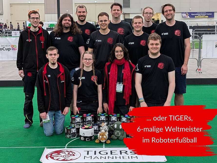 Eine Gruppe von elf Personen steht gemeinsam auf einem grünen Roboterfußballfeld. Die meisten tragen schwarze TIGERs-Shirts. Vor der Gruppe liegen mehrere kleine Roboter und Maskottchen. Im unteren Bildbereich ist ein Banner mit der Aufschrift „TIGERS Mannheim“ zu sehen. Rechts steht: … oder TIGERs, 6-malige Weltmeister im Roboterfußball