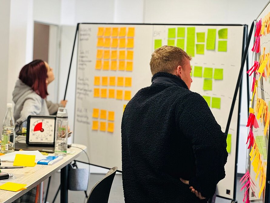 Fokus auf einen Studenten vor einem Whiteboard mit Post-its; im Vordergrund ein Time-Timer zur Zeitsteuerung des Workshops.