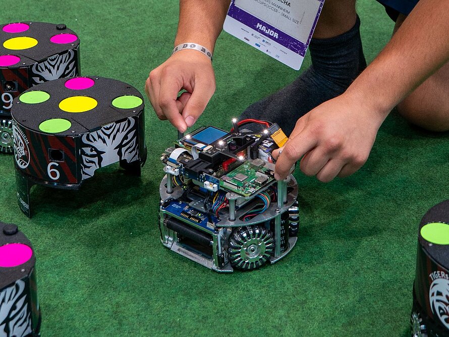 5 Fußballroboter auf grünem Feld, der mittlere davon ohne Hülle. Ein Spieler der TIGERs nur mit Händen zu sehen, die an dem Roboter hantieren. 