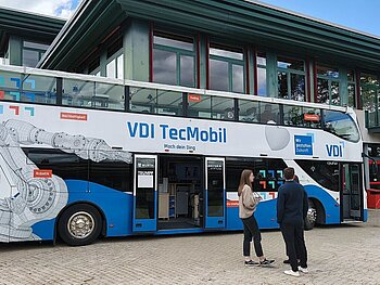 Blick auf das TecMobil von der Seite, davor stehen 3 Personen im Alter von ca. 20 Jahren, die sich unterhalten. Das TecMobil ist ein doppelstockiger Bus in blau-weiß, auf dem ein Roboterarm sowie Schlagworte wie Nachhaltigkeit,  Coding, Robotik und Konstruktion abgebildet sind. 