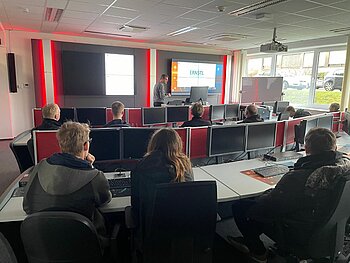 Blick von den hinteren Reihen des Cyber Security Labors der DHBW Mannheim, das mit vielen Monitoren ausgestattet ist und in dem ein Vortrag von einem Professor gehalten wird. 