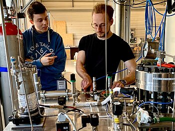 Zwei Studierende von vorne zu sehen. Sie arbeiten an einem Tisch mit vielfältigen technischen Vorrichtungen, darunter ein Gas-Behälter für Wasserstoff. 