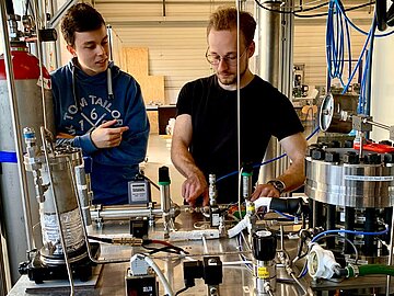 Zwei Studierende von vorne zu sehen. Sie arbeiten an einem Tisch mit vielfältigen technischen Vorrichtungen, darunter ein Gas-Behälter für Wasserstoff. 