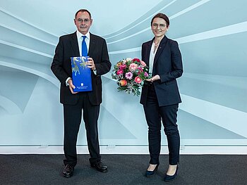 Ein Mann und eine Frau, beide in schwarzem Anzug stehen vor einem grauen Hintergrund-Aufsteller mit dynamischen Linien, sie mit Blumen er mit einer Urkunde