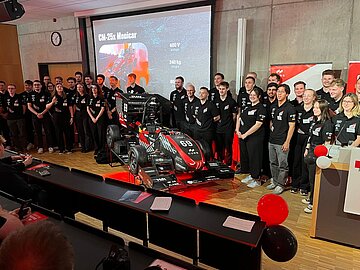 Gruppenbild mit 35 Team-Mitgliedern und dem Rennwagen in der Mitte, dahinter an der Wand die Präsentation mit wichtigsten Eckdaten zum Auto. Die Reihen des Hörsaals sind mit roten und schwarzen Luftballons geschmückt. 