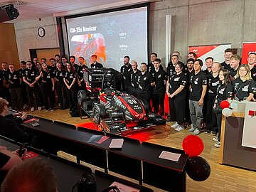 Gruppenbild mit 35 Team-Mitgliedern und dem Rennwagen in der Mitte, dahinter an der Wand die Präsentation mit wichtigsten Eckdaten zum Auto. Die Reihen des Hörsaals sind mit roten und schwarzen Luftballons geschmückt. 