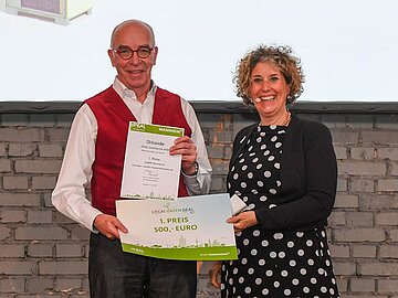 Porträt von zwei Menschen. Links ein Herr, der eine Urkunde von einer rechts neben ihm stehenden Dame erhält. Beide Lachen in die Kamera. 
