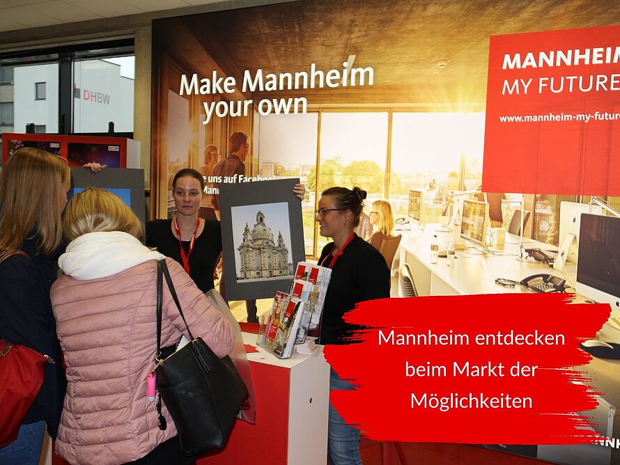 Studierende informieren sich an einem Ausstellerstand beim Markt der Möglichkeiten über Angebote in Mannheim. Im Hintergrund steht „Make Mannheim your own“. Der eingeblendete Text lautet: Mannheim entdecken beim Markt der Möglichkeiten.