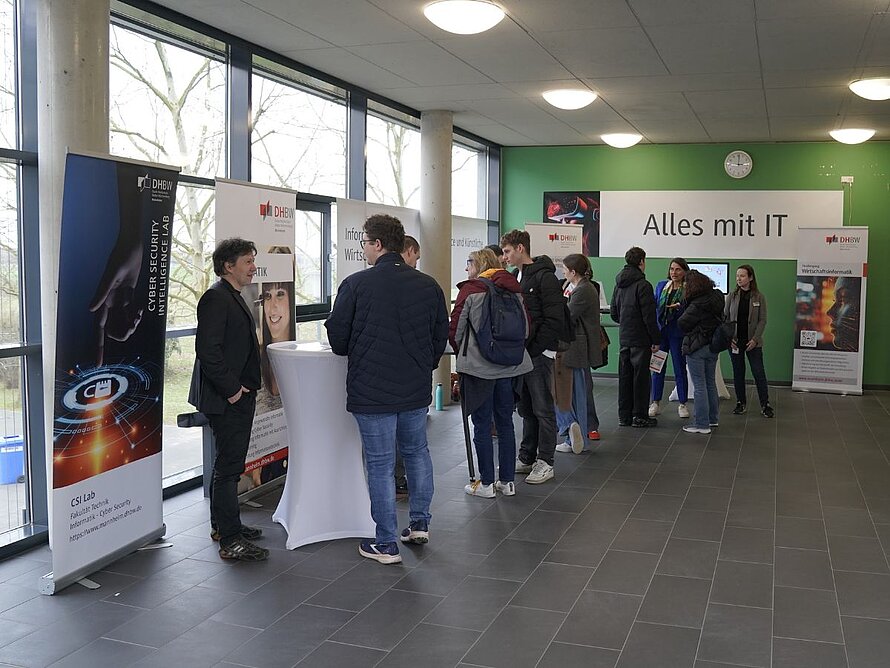 Gespräch von mehreren Menschen am Stand der Informatik, im Hintergrund Plakate an der Wand mit den Namen der Studiengänge