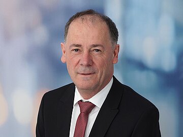 Porträt Prof. Dr. Ludwig Bauer lächelnd, mit schwarem Anzug und dunkelroter Krawatte vor einem Hintergrund in unterschiedlichen Blautönen.