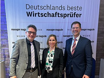 Gruppenbild mit zwei Männern und einer Frau in der Mitte - im Hintergrund ein Banner mit dem Titel "Deutschlands beste Wirtschaftsprüfer", darunter Sponsorenlogos. 