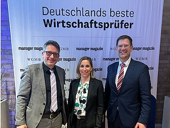 Gruppenbild mit zwei Männern und einer Frau in der Mitte - im Hintergrund ein Banner mit dem Titel "Deutschlands beste Wirtschaftsprüfer", darunter Sponsorenlogos. 