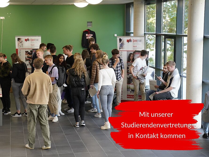 Studierende kommen an einem Stand der Studierendenvertretung der DHBW Mannheim miteinander ins Gespräch. Informationsmaterial und Infostände der StuV sind im Hintergrund zu sehen. Der eingeblendete Text lautet: Mit unserer Studierendenvertretung in Kontakt kommen.