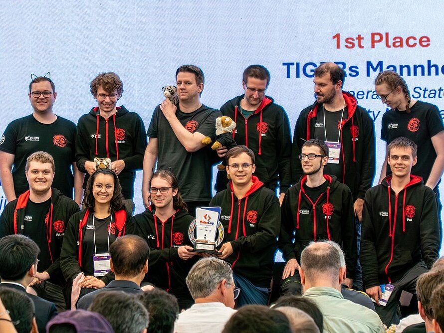Gruppenbild mit 12 Personen in schwarz-roten Team-Hoodies stehen in 2 Reihen, ein Student in der vorderen Reihe hält die Siegertrophäe. Auf einer weißen Leinwand dahinter steht der Team-Name samt Platzierung. 