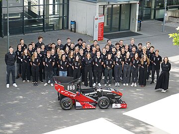 Gruppenbild aus der Vogelperspektive fotografiert mit über 55 Studierenden in schwarzen CURE-Poloshirts auf dem Außengelände der DHBW Mannheim, davor der neue Rennwagen.
