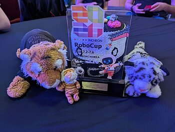 Auf dem Tisch steht die Trophäe des RoboCups, neben der Trophäe liegen zwei Plüschtiger und ein Plüschleopard