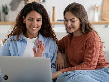 Zwei Frauen sitzen lächelnd nebeneinander vor einem Laptop