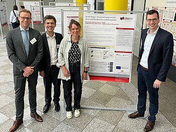 Gruppe bestehend aus drei Männern und einer Frau an einem Poster, auf dem das Forschungsprojekt vorgestellt wird. Im Hintergrund befinden sich noch viele weitere Pinnwände mit Projektbeschreibungen. 