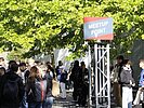 Menschen stehen auf einem Außenbereich des Campus an einem Treffpunkt zusammen. Über dem Treffpunkt hängt ein Schild mit der Aufschrift „MEETUP POINT“. Im Hintergrund sind Bäume und weitere Studierende zu sehen