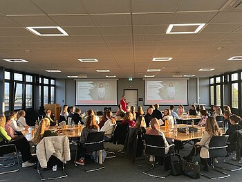 Eine Zuhörerschaft von mehr als 30 Personen an 4 Gruppentischen blickt auf die Referentin und deren Powerpoint-Präsentation. 