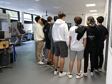 9 Schüler*innen im Oberstufenalter von hinten zu sehen, die im Halbkreis im Maschinenbaulabor der DHBW Mannheim stehen und sich die Funktionsweise der Maschinen erklären lassen. 