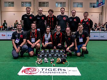 Gruppenbild mit 12 Menschen in 2 Reihen, davor 11 Fußball-Roboter auf dem grünen Teppich des Spielfelds sowie die Flagge des Teams TIGERs Mannheim.