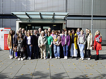 Gruppenbild im herbstlichen Sonnenschein mit 40 Personen auf dem Außengelände der DHBW Mannheim; hinter den in Jacken gehüllten Personen der Eingang zu Gebäude D mit heruntergefahrenen Jalousien.