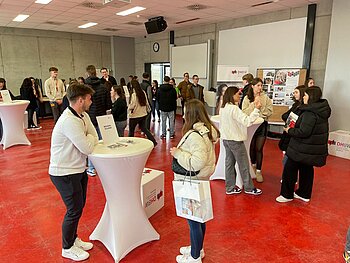 Raum mit Stehtischen zu unterschiedlichen Studiengängen der DHBW Mannheim, an denen Studierende und Studieninteressierte sich austauschen.