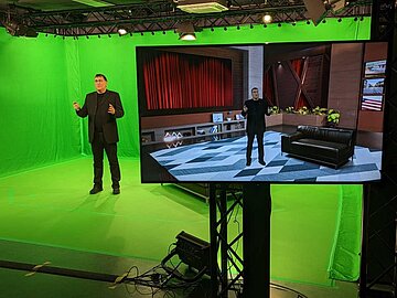 Prof. Dr. Dinkel steht im Greenscreen-Labor, rechts voran steht ein Bildschirm, auf dem er vor einem virtuellen Hintergrund gezeigt wird.