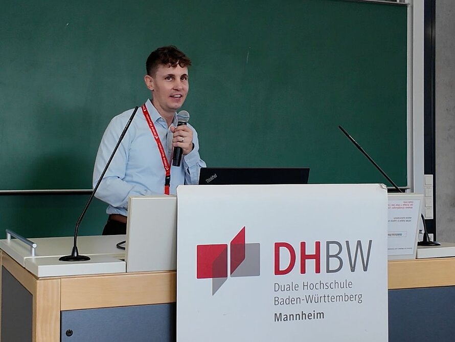 Mann mit Mikrofon am Redepult spricht zum Plenum