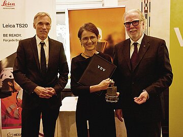 Gruppenbild mit drei lachenden Personen, in der Mitte eine Frau, die eine Urkunde des Verbands Deutscher Vermessungsingenieure in der Hand hält. 